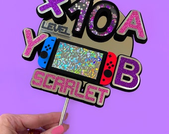 Könnte beinhalten: Ein rosa und lilafarbener Tortendekor mit einem Videospiel-Controller, den Worten "Level 10A", "Scarlet" und "Y" sowie einem glitzernden lila "B".
