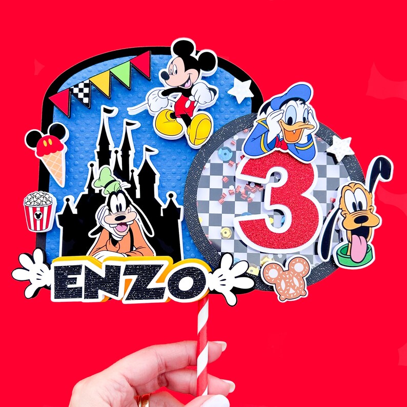 May include: A colorful cake topper featuring Mickey Mouse, Donald Duck, Goofy, and the Disney castle. The topper also includes a red number 3, a checkered pattern, and the name "ENZO".