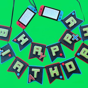 Puede incluir: Una pancarta negra y verde con las palabras "Feliz cumpleaños" escritas en letras de colores. La pancarta está decorada con imágenes de mandos de videojuegos.