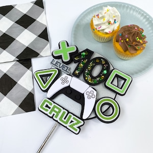 May include: Green and white cake topper with a video game controller, a plus sign, the number 10 with green glitter, a triangle, a square, a circle, and the word "LEVEL" and "CRUZ".