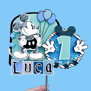 May include: A blue and black Mickey Mouse cake topper with a number 1 and the name Luca. The topper features a cartoon Mickey Mouse with black and white outlines, blue balloons, and a blue and white striped background.