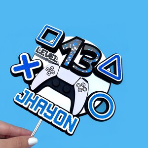 May include: A blue and white cake topper with a video game controller, a blue and white 'X' symbol, a blue and white triangle, a blue and white circle, a blue and white square, and the number '13' in black and white with blue glitter. The text 'Level' and 'Jhayon' are also included in blue and white.