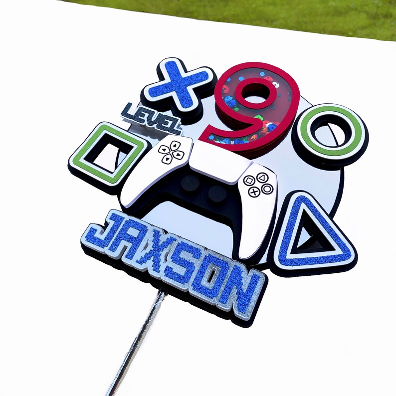 May include: A blue and silver cake topper with the name Jaxson, a video game controller, a blue triangle, a green square, a red number 9 with glitter, a blue X, and the words "Level" on it.
