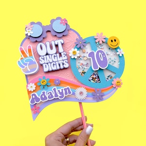 May include: A pink and blue cake topper with a peace sign, rainbow, and the text "Out Single Digits" and "Adalyn".