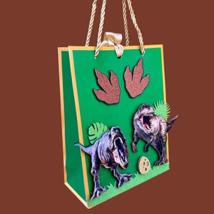 Pode incluir: Uma sacola de presente verde com acabamentos e al&ccedil;as douradas, com ilustra&ccedil;&otilde;es de dinossauros e pegadas brilhantes. A sacola tem um la&ccedil;o de fita e apresenta dinossauros de desenho animado, incluindo um T-Rex.