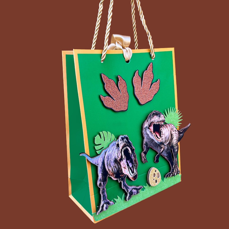 Pode incluir: Uma sacola de presente verde com acabamentos e al&ccedil;as douradas, com ilustra&ccedil;&otilde;es de dinossauros e pegadas brilhantes. A sacola tem um la&ccedil;o de fita e apresenta dinossauros de desenho animado, incluindo um T-Rex.