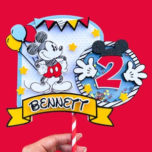 May include: A Mickey Mouse themed cake topper with a cartoon Mickey Mouse holding balloons, a red number 2 with Mickey Mouse hands, and a yellow banner with the name "BENNETT".