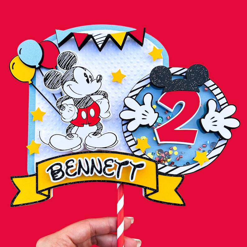 May include: A Mickey Mouse themed cake topper with a cartoon Mickey Mouse holding balloons, a red number 2 with Mickey Mouse hands, and a yellow banner with the name "BENNETT".