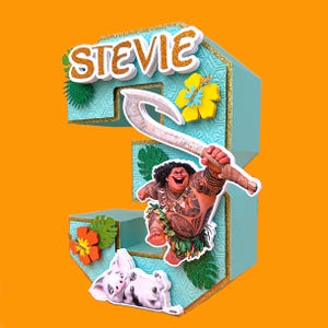 Puede incluir: Un adorno para tarta con el n&uacute;mero 3 en color turquesa con el nombre "Stevie" en purpurina dorada. El adorno presenta una imagen de dibujos animados de Maui de Moana sosteniendo su anzuelo, junto con Pua y una flor de hibisco amarilla.
