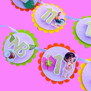 Puede incluir: Una pancarta festiva con fondo rosa, con recortes circulares con patrones en relieve y bordes festoneados en varios colores. Cada recorte muestra un n&uacute;mero, un personaje de dibujos animados y elementos tropicales como cocos y flores. Los n&uacute;meros est&aacute;n delineados en purpurina dorada.