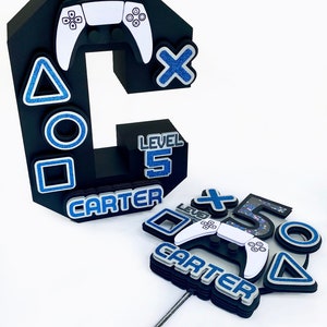 May include: A black and white cake topper with a video game controller, blue and silver geometric shapes, and the text "CARTER" and "LEVEL 5".