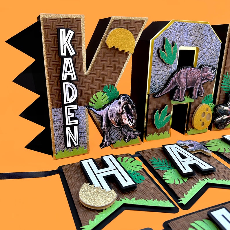 Pode incluir: Uma faixa de anivers&aacute;rio com tema de dinossauro com o nome "KADEN" em letras grandes 3D. As letras s&atilde;o marrons com um padr&atilde;o texturizado, acentuadas com glitter dourado e ilustra&ccedil;&otilde;es de dinossauros. A faixa tamb&eacute;m diz "HAPPY".