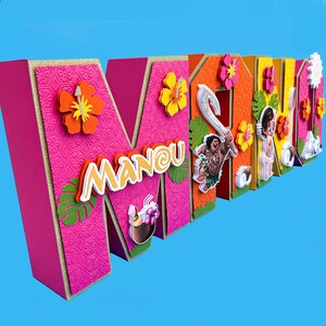 Puede incluir: Un letrero de nombre de cart&oacute;n rosa, naranja y amarillo con el nombre "MANOU" en letras blancas. El letrero est&aacute; decorado con flores, hojas y un personaje de dibujos animados de la pel&iacute;cula Moana.