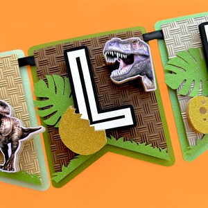 Pode incluir: Uma faixa com tema de dinossauro com detalhes em verde, marrom e dourado. A faixa apresenta um fundo texturizado, uma grande letra "L", ilustra&ccedil;&otilde;es de dinossauros e folhas decorativas. A faixa &eacute; projetada para um anivers&aacute;rio ou festa.