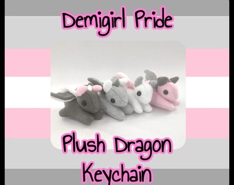 Może przedstawiać: Wyświetlane są cztery pluszowe smocze breloczki w odcieniach szarości, bieli i różu. Tekst "Demigirl Pride" i "Plush Dragon Keychain" jest napisany różowym pismem. Tło przedstawia flagę Demigirl Pride.