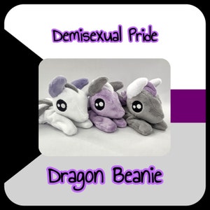May include: Three plush dragon beanies in white, purple, and gray colors. The beanies are arranged in a row and have black eyes. The text "Demisexual Pride" is above the beanies and "Dragon Beanie" is below.