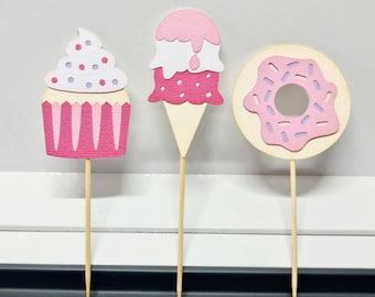 May include: Three pink and white paper cupcake, ice cream cone, and donut toppers on wooden sticks. The cupcake has purple sprinkles, the ice cream cone has white and pink sprinkles, and the donut has pink sprinkles.