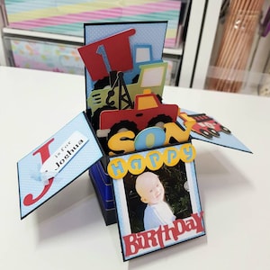 Op de afbeelding: Een handgemaakte pop-up verjaardagskaart met een babyfoto. De kaart heeft een rood en blauw vrachtwagenthema met de woorden "Happy" en "Birthday". De kaart bevat ook de tekst "J is for Joshua".