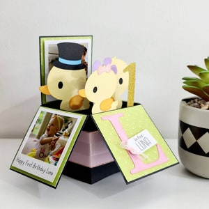 Peut inclure: Une boîte pop-up verte et rose avec une photo d'une petite fille et un canard portant un chapeau haut de forme. La boîte a un fond vert à pois blancs et le texte "Happy First Birthday Luna". La boîte a également une lettre L rose avec le texte "L is for Luna" sur une étiquette blanche.