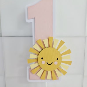 May include: Pink number one cake topper with a yellow sun with a smiling face. The sun has pink cheeks.