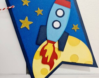 May include: A blue banner with a rocket ship and stars. The rocket is light blue with red and white accents, and the banner has a yellow moon shape at the bottom. Gold glitter stars are scattered across the blue background.