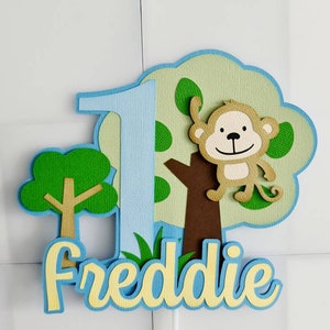Peut inclure: Décoration de gâteau en papier superposé avec un singe, un arbre et le chiffre 1. Le design comprend le nom "Freddie" dans une police ludique, avec des couleurs bleu, vert, marron et jaune. La décoration est conçue pour une fête d'anniversaire.