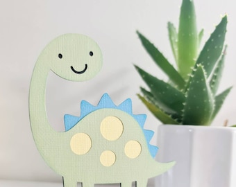 May include: A light green dinosaur cake topper with a smiling face, blue spikes, and yellow spots. The dinosaur is made of paper and is on a white stick. A green plant in a white pot is in the background.