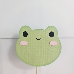 May include: A light green frog-shaped paper cutout with black eyes and pink cheeks. The frog has a smiling mouth and is attached to a wooden stick. The background is white.