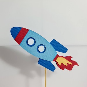 May include: A blue and red rocket ship cake topper with a yellow flame. The rocket ship has two white circles for windows.