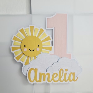 May include: A pink number one cake topper with a yellow sun and white cloud. The sun has a smiling face and the cloud has the name Amelia written on it.