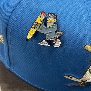 May include: A blue baseball cap with a gray and yellow enamel pin of a cartoon bird holding a pencil. The bird is wearing a blue jacket and a black hat.