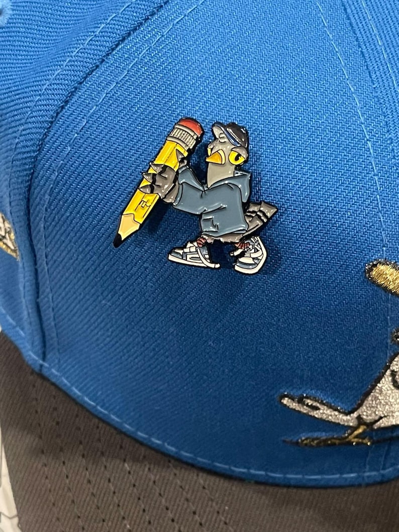 May include: A blue baseball cap with a gray and yellow enamel pin of a cartoon bird holding a pencil. The bird is wearing a blue jacket and a black hat.