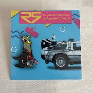 May include: A pair of enamel pins featuring a cartoon pigeon wearing a hat and a DeLorean time machine. The pins are on a blue background with a pink and yellow design. Etsy: RobSmidDesigns IG: Rob_Smid_Designs