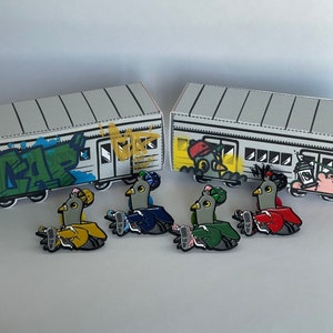 May include: Four enamel pins featuring cartoon pigeons in different colors, including blue, green, red, and yellow. The pins are set against a backdrop of two miniature model train cars with graffiti.
