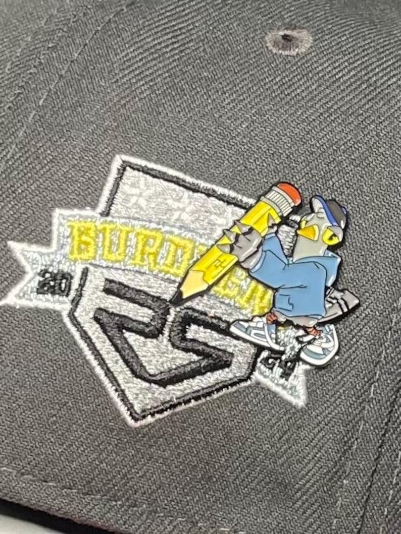 May include: A gray baseball cap with a silver and black embroidered patch featuring a cartoon character holding a yellow pencil. The patch says "BURDMAN 2023".