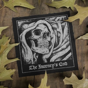 May include: Black and white embroidered patch with a skull in a hooded cloak. The text "The Journey's End" is below the skull.