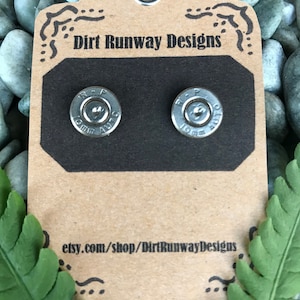 Puede incluir: Pendientes plateados con forma de casquillos de bala, presentados en una etiqueta marrón con el texto "Dirt Runway Designs" y "etsy.com/shop/DirtRunwayDesigns". Los pendientes se colocan sobre un fondo de piedras grises y hojas verdes.