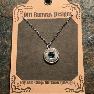 May include: A silver-toned necklace featuring a pendant made from a .45 AUTO Winchester bullet casing. The bullet casing holds a small, round, emerald-green gemstone. The necklace hangs on a delicate chain, displayed on a brown card with the text "Dirt Runway Designs".
