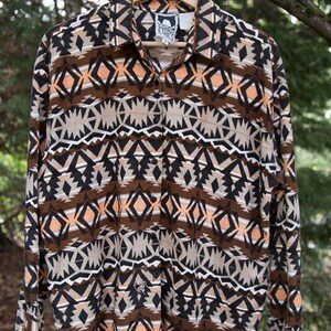 Puede incluir: Una camisa de manga larga con botones y un patrón geométrico en tonos de marrón, negro, blanco y naranja. La camisa tiene cuello y cierre de botones en la parte delantera. La etiqueta de la camisa dice "Ethics".