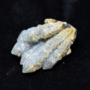Puede incluir: Un racimo de cristales de cuarzo gris pálido translúcido con una superficie rugosa y texturizada. La formación cristalina presenta puntas dentadas y está cubierta con un depósito mineral fino, arenoso y de color marrón dorado.