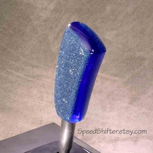 Può includere: Un pomello del cambio blu glitterato con una finitura lucida e liscia. La parte superiore è ricoperta di glitter scintillanti, mentre i lati sono di un blu intenso e lucido. Il testo "SpeedShifter.etsy.com" è visibile.