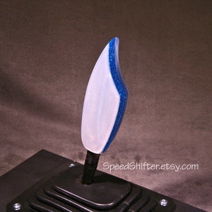 Op de afbeelding: Een aangepaste auto versnellingspookknop met een wit en glinsterend blauw ontwerp. De versnellingspookknop is gemonteerd op een zwarte basis. De tekst "SpeedShifter.etsy.com" is zichtbaar.