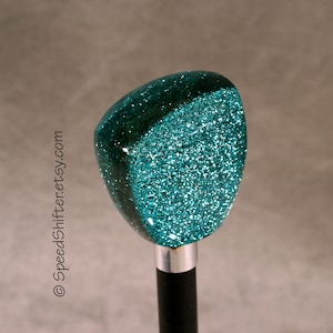 May include: A teal glitter shift knob with a silver base and black shaft. The top of the knob is a rounded triangle shape. The glitter is embedded in a clear resin. The text "SpeedShifter.etsy.com" is visible.