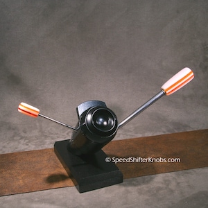 May include: A black speed shifter knob with two silver arms and white and orange striped handles. The knob is mounted on a black base. The website SpeedShifterKnobs.com is visible on the image.