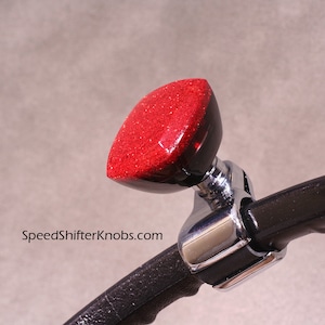 May include: A red glitter shift knob with a chrome base is attached to a black steering wheel. The text "SpeedShifterKnobs.com" is visible on the image. The shift knob has a unique, eye-catching design.