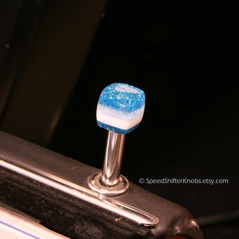 May include: A custom car door lock knob with a blue glitter top, a white stripe, and a silver metal stem. The knob is a unique accessory for car interiors.
