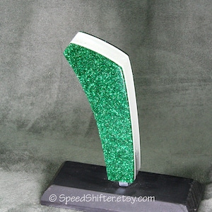 May include: A custom car shift knob with a green glitter finish and white trim. The shift knob is mounted on a black base. The Etsy shop name SpeedShifter.etsy.com is printed on the base.
