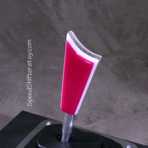 Op de afbeelding: Een aangepaste auto versnellingspookknop met een fuchsia en wit ontwerp. De knop heeft een zilveren glitterrand en is gemonteerd op een zwarte basis. De tekst "SpeedShifter.etsy.com" staat op de zijkant.