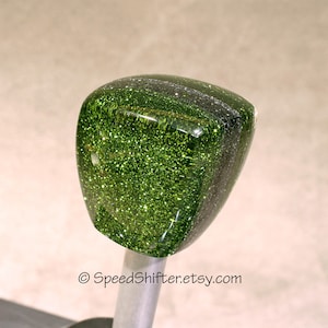 May include: A green glitter-filled shift knob with a rounded, rectangular shape. The knob has a glossy finish and is mounted on a silver-colored metal rod. The Etsy shop name "SpeedShifter.etsy.com" is printed at the bottom.