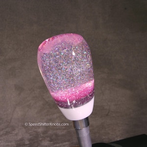 May include: A gear shift knob featuring a clear, rounded top filled with iridescent silver glitter, transitioning into a layer of bright pink glitter and a dark purple glitter base, set atop a solid white cylindrical mount with a metal stem.
